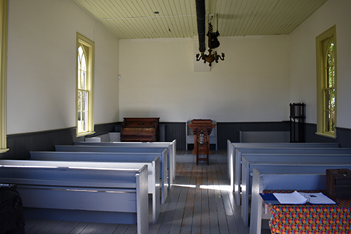 Intérieur de la Pioneer Church au Musée Josiah Henson de l’histoire afro-canadienne, montrant des rangées de bancs en bois simples faisant face à une chaire et à l’orgue original au fond de la salle. La lumière du soleil traverse les hautes fenêtres des deux côtés.