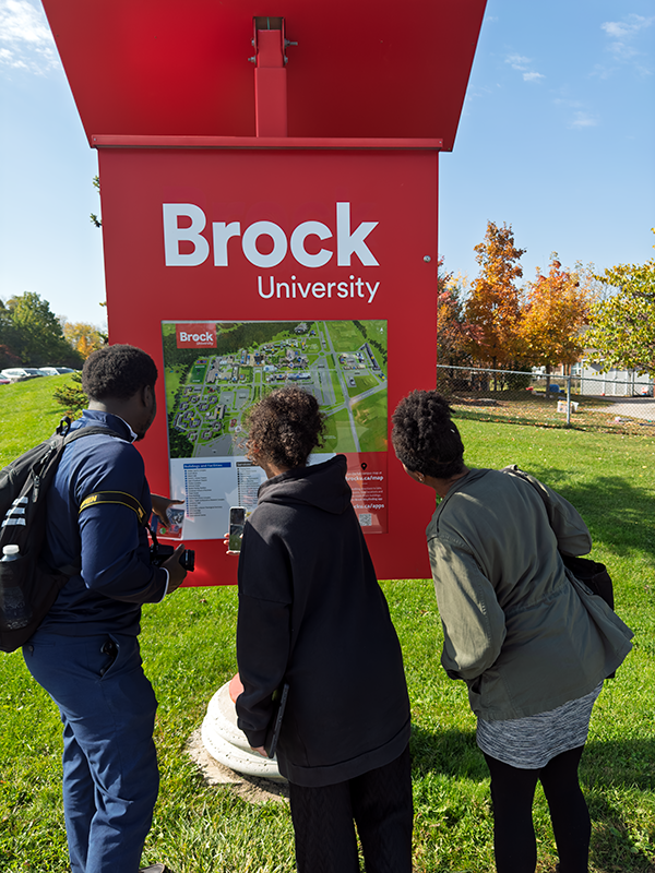 Alex Allasra, Gabriela Sealy et la Dre Émilie Andrée Roumer Jabouin regardent une grande carte d’orientation rouge à l’Université Brock. Des arbres d’automne et un ciel lumineux apparaissent à l’arrière-plan. Photo de Blessing Ogunyemi.