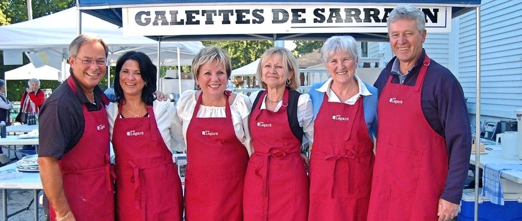 Plan d’ensemble de six personnes portant des tabliers rouges se tiennent debout devant une tente pour la vente de galettes de sarrasin. 