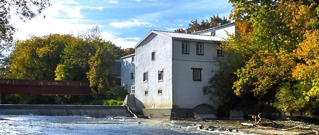 Un bâtiment blanc à toit en pente. Un pont en bois surplombe la digue de la rivière qui se trouve à l’avant-plan.