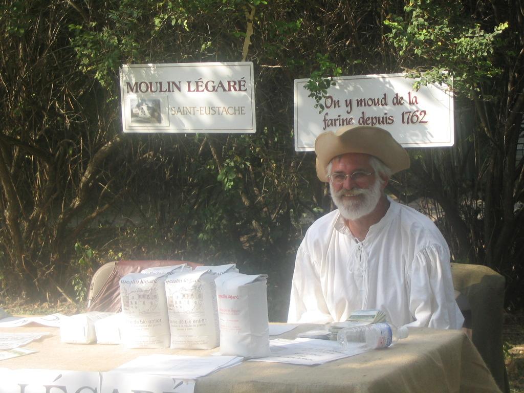 Pierre-Paul Renaud portant une barbe blanche, un tricorne beige et une chemise blanche, assis à l'extérieur, derrière une table de vente de farine.	