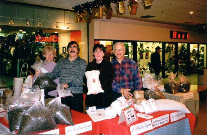 Quatre bénévoles sont debout derrière une table et tiennent des sacs de farine dans un centre commercial décoré pour Noël.