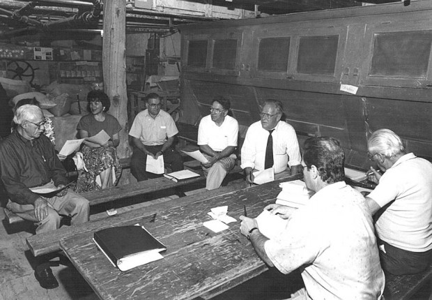 Photo noir et blanc de sept membres du conseil d’administration assis en cercle sur des bancs en bois et tenant des feuilles.
