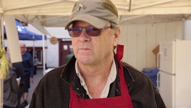 Plan rapproché de Luc Raby portant une casquette kaki, des verres fumés et un tablier rouge.