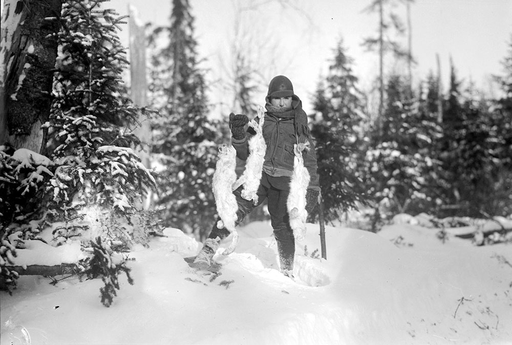 Photographie noir et blanc d'un enfant en raquettes qui tient trois lièvres, des prises de chasse, en forêt.