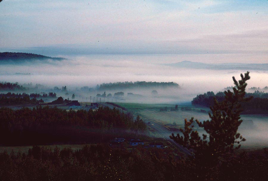 Vue d'un paysage embrumé. Au centre, un village dont on distingue quelques édifices, traversé par une route.