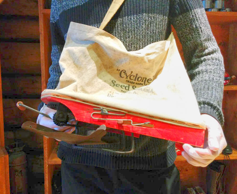 Un animateur de musée tient en bandoulière un semoir en bois rouge avec distributeur rotatif de métal et muni d'une pochette de coton.