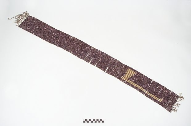 Ceinture de perles tissées, de forme rectangulaire allongée, aux teintes mauves et blanches. On y décerne la forme d'une hache comme décoration.