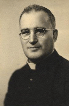 Photographie noir et blanc : un portrait officiel d'un homme en habit de curé, portant des lunettes.