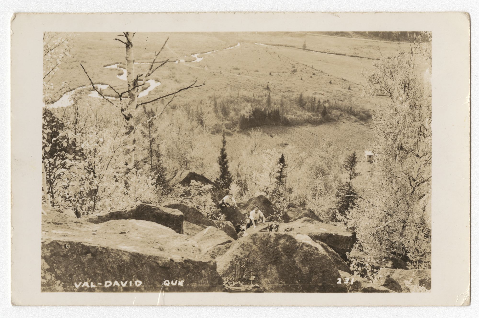 Carte postale prise en plongée depuis un sommet; deux hommes grimpent un amoncellement de rochers. En arrière-plan une partie de la vallée de Val-David. 