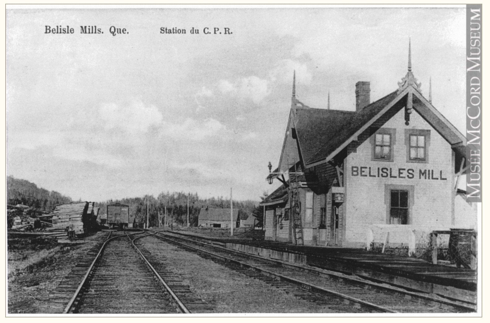 Carte postale ancienne illustrant la gare ferroviaire de Bélisle’s Mills; on y remarque l’édifice, les voies ferrées, les tas de bois prêts pour l’expédition.