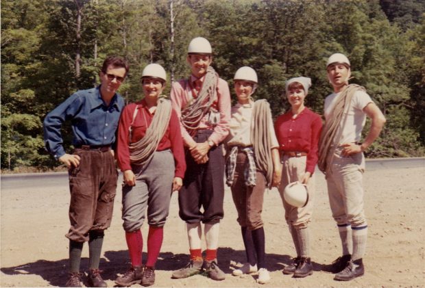 Un groupe de six grimpeurs et grimpeuses, sur un chemin devant une forêt, avec leurs casques et des cordes.