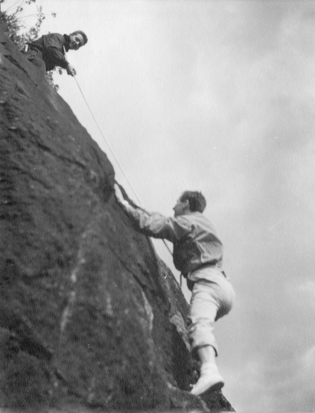Deux grimpeurs, l’un au sommet d’un rocher retenant une corde à laquelle est accroché un second grimpeur en cours d’escalade.