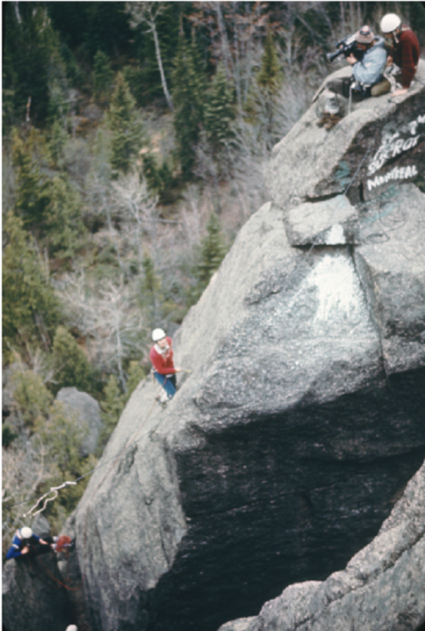 Groupe escaladant un rocher placé haut dans la forêt. Un cameraman et un teneur de corde sont au sommet; une femme grimpe le rocher.