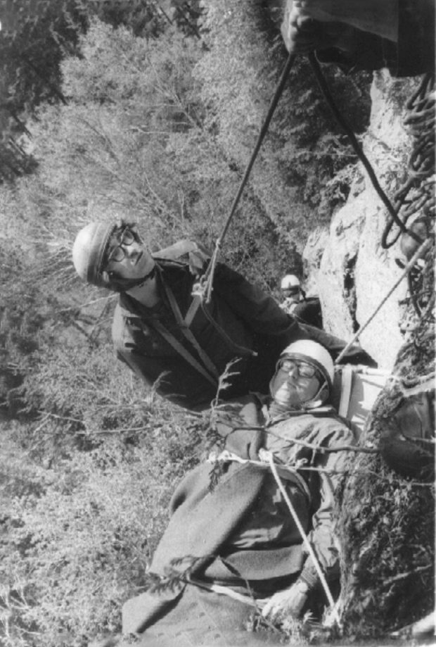 Vue en plongée de deux hommes en pleine paroi; un homme est étendu dans une civière et l’autre l’aide à descendre la paroi à l’aide de cordages.