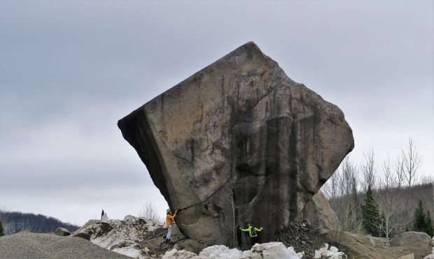 Un immense bloc de roche de forme carré d’environ 12 mètres de hauteur avec à sa base deux personnes qui l’inspectent.