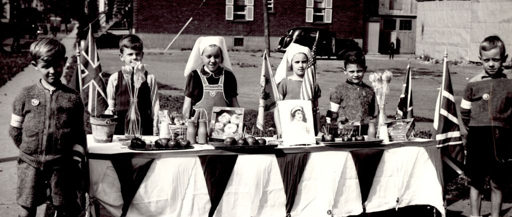 Portrait de six enfants, deux filles au centre et deux garçons de chaque côté, derrière une table remplie de nourriture, quatre drapeaux de l'Union Jack et deux vases de fleurs. Les jeunes filles portent un couvre-chef de type religieuse alors que les garçons portent une épinglette sur leur veste. 