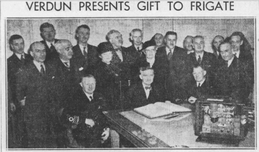 Photographie en noir et blanc titrée « Verdun presents gift to frigate ». Dans une salle, au bout d’une table sur laquelle est posé un projecteur et un grand livre ouvert se trouvent 3 personnes assises et 16 debout. 