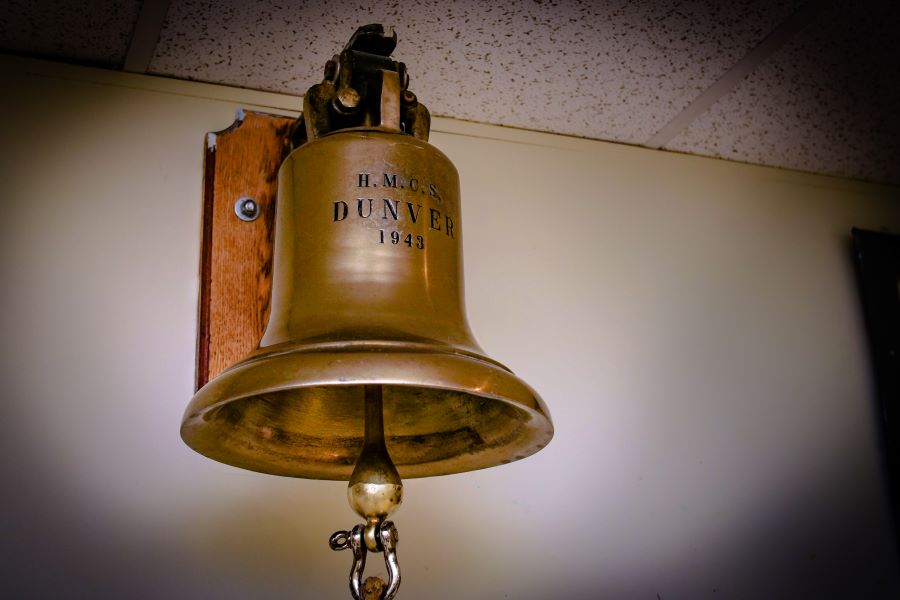 Photographie couleur d’une cloche accrochée sur un mur d’un local. L’inscription HMCS DUNVER 1943 est gravée sur la cloche de couleur dorée.