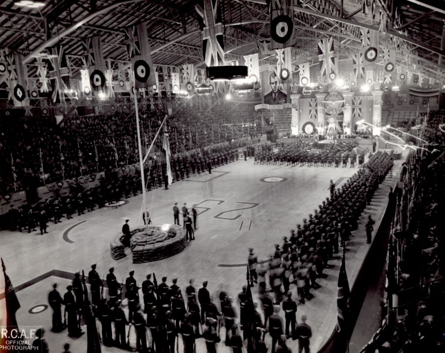 Photographie noir et blanc. Une scène est installée au bout d’une patinoire dans un aréna. Des rangs de militaires devant et sur les côtés. Des drapeaux d'aviation et d’Union Jack sont suspendus au plafond. Les estrades sont remplies de gens.