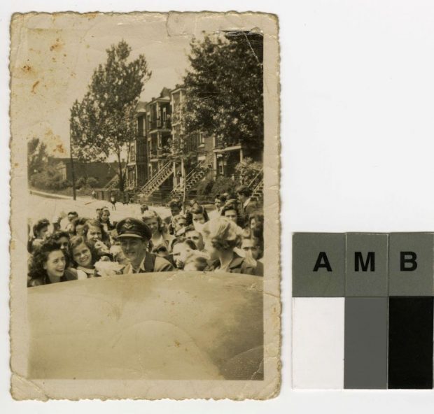 Photographie noir et blanc d’un jeune homme souriant en uniforme prêt à embarquer dans une voiture. Il est entouré d’un groupe d’environ 20 jeunes, filles et garçons. En arrière-plan: une rue, des bâtiments et des arbres.