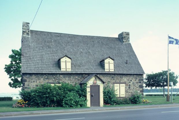 Photographie couleur d’une maison ancienne de style breton qui comporte une porte d’entrée au centre, deux fenêtres au rez-de-chaussée et deux lucarnes au grenier. L’emblème de la Légion canadienne est installé au-dessus de la porte. Un drapeau québécois flotte à droite de la maison. Le fleuve Saint-Laurent est visible en arrière-plan.