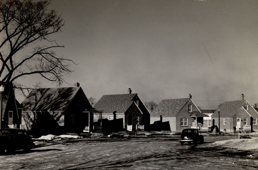 Photographie noir et blanc de quatre petites maisons de type cottage dans une rue. Deux voitures stationnées dans la rue sont visibles à l’avant. À gauche, la moitié d’un grand arbre. 