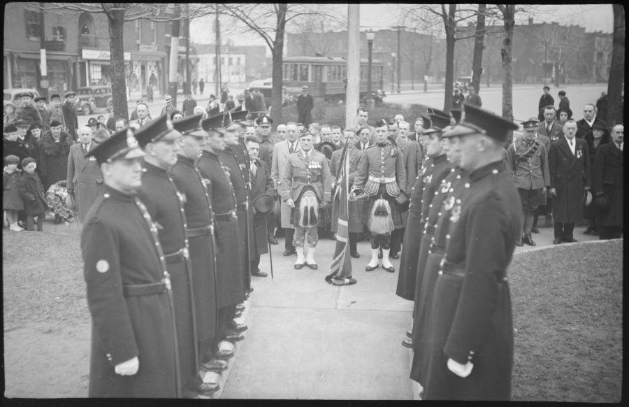 Photographie noir et blanc. Au centre, 12 militaires en uniforme forme une garde d’honneur devant deux militaires en kilts et un homme en civil, dont un portant un drapeau. Derrière eux se trouvent une cinquantaine de dignitaires et spectateurs. À l'arrière-plan, quelques arbres, la rue et des bâtiments.