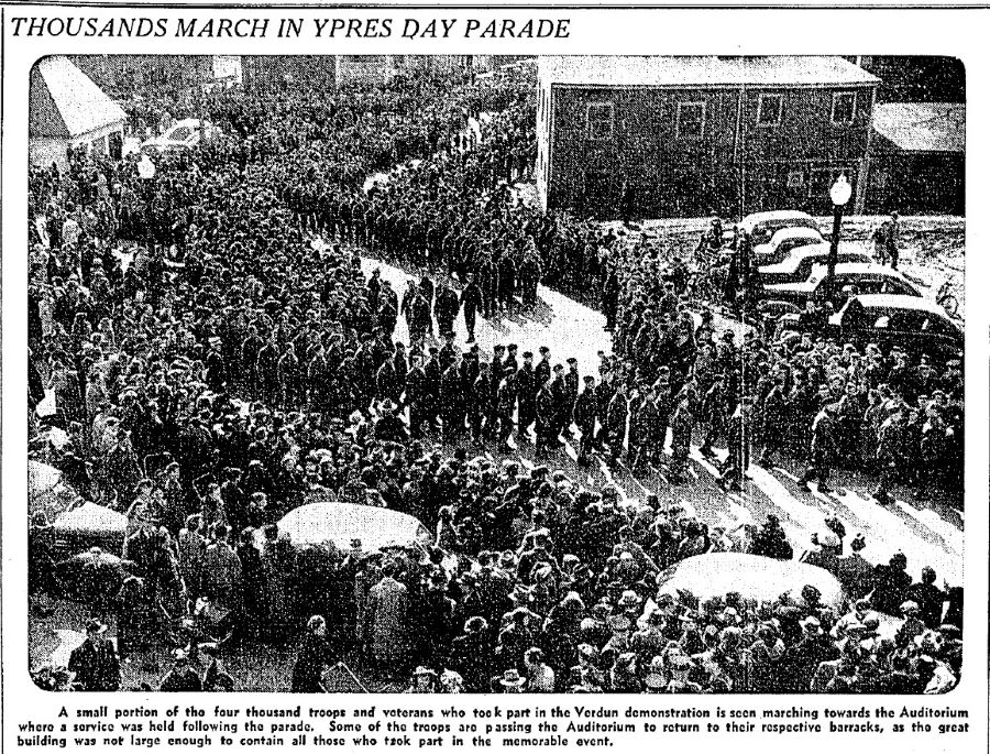 Article de journal, noir et blanc, titré « Thousands March in Ypres Day Parade ». En dessous une grande photographie de centaines de personnes marchant dans une rue, sur les côtés des voitures stationnées entourées de spectateurs. Au bas se trouve un texte de 65 mots. 