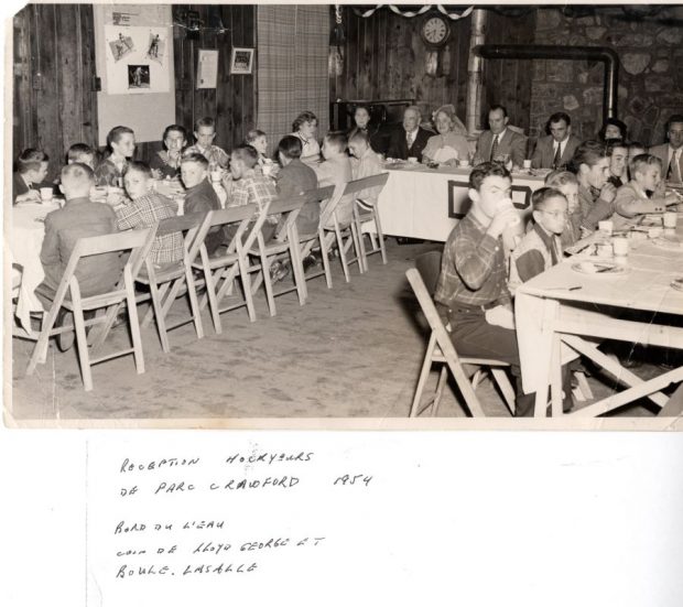  Photographie noire et blanc sur laquelle nous pouvons voir la présence de sept adultes qui sont assis à une table. Deux groupes d’enfants prennent place autour de deux autres tables, dont 14 enfants à gauche et sept autres à droite  de celle qui réunit les adultes au centre.  En arrière-plan se trouvent des murs en pierre, une horloge et des cadres.