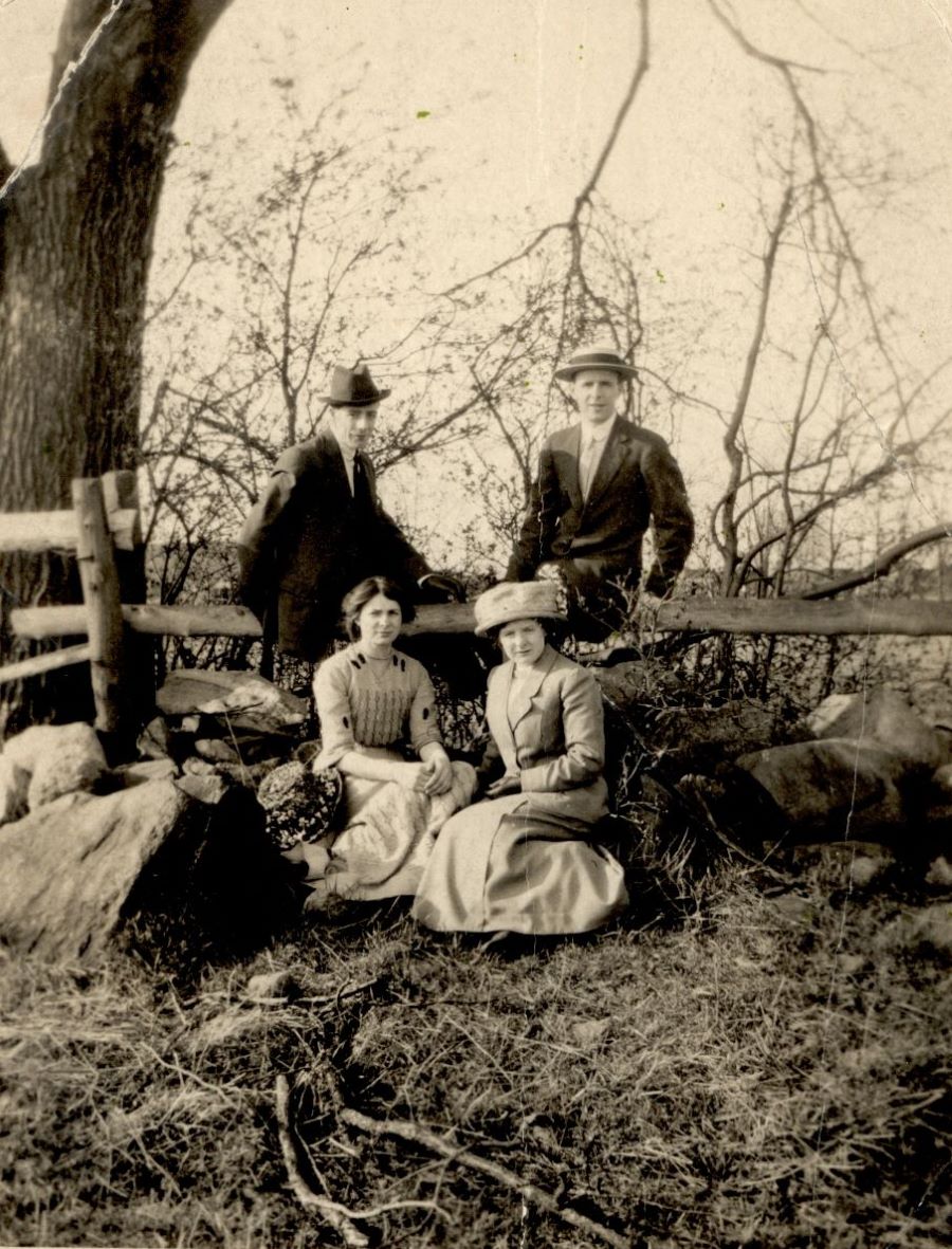 Photographie en noir et blanc d’un groupe de quatre adultes élégamment vêtus: deux hommes debout appuyés sur une clôture de perches en bois et devant, deux femmes assises sur l’herbe. À l’arrière-plan, un arbre et un champ.