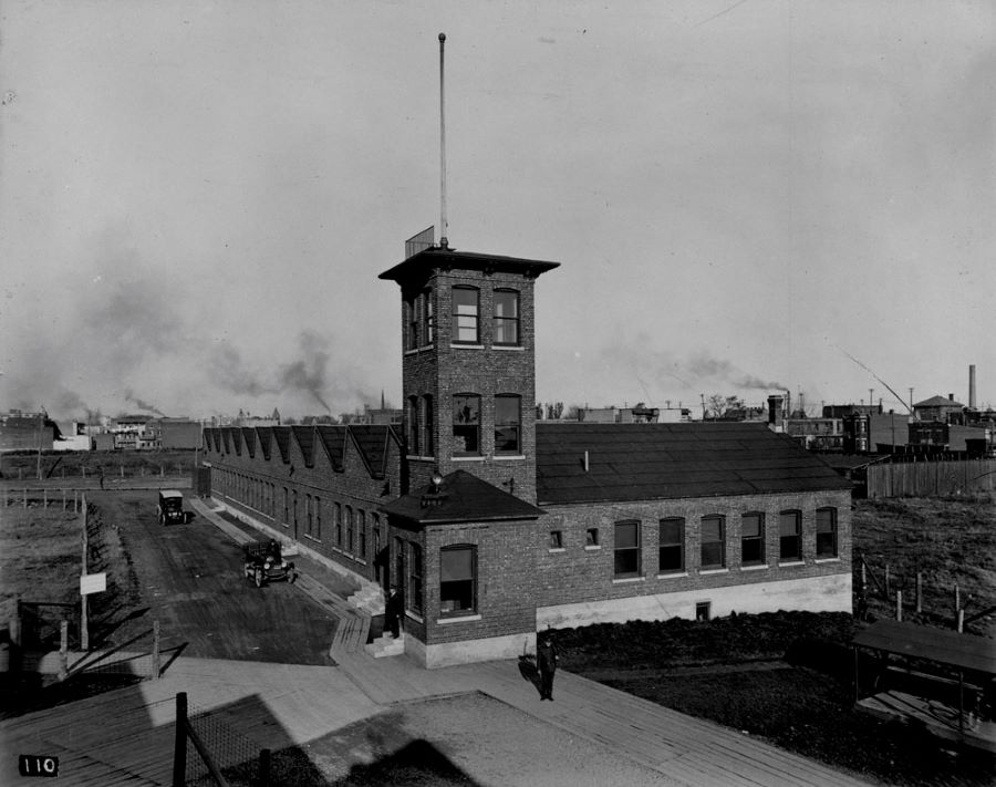 Photographie en noir et blanc d’un bâtiment industriel avec une tour d’angle, plusieurs fenêtres sur les façades et un toit en dent de scie. À l'arrière-plan, de la fumée sort des cheminées d'industries.