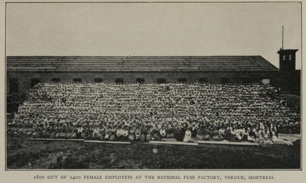 Photographie en noir et blanc montrant 1 800 femmes assises en une vingtaine de rangées sur une petite colline devant l'usine de munitions.