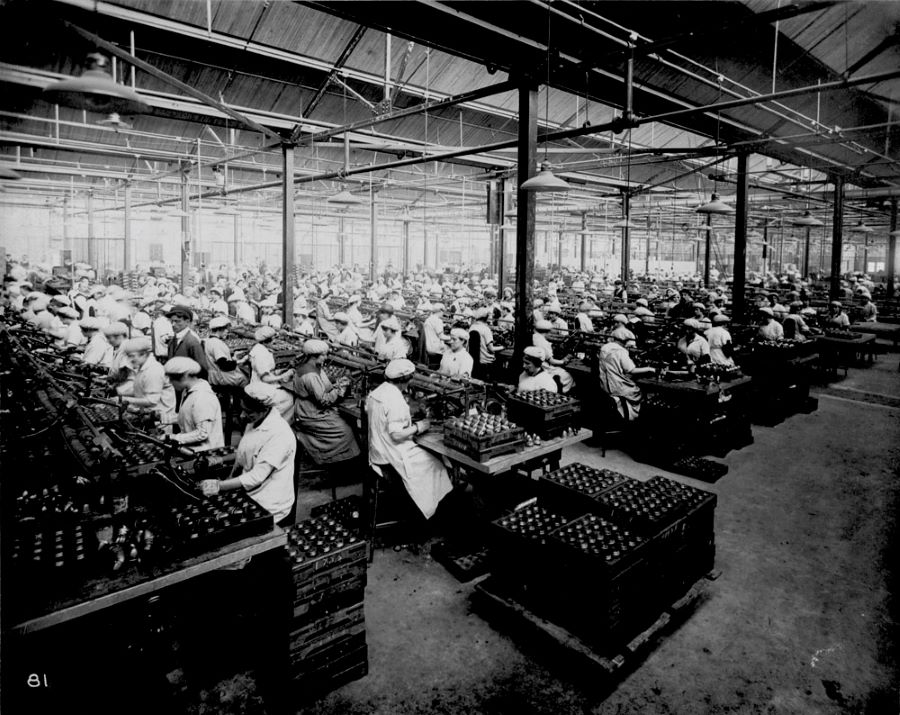 Photographie en noir et blanc de plusieurs rangées d’ouvrières au travail à l'intérieur de l'usine. Elles sont assises près les unes des autres à leur poste de travail. Dans l'une des rangées, un homme surveille le travail.