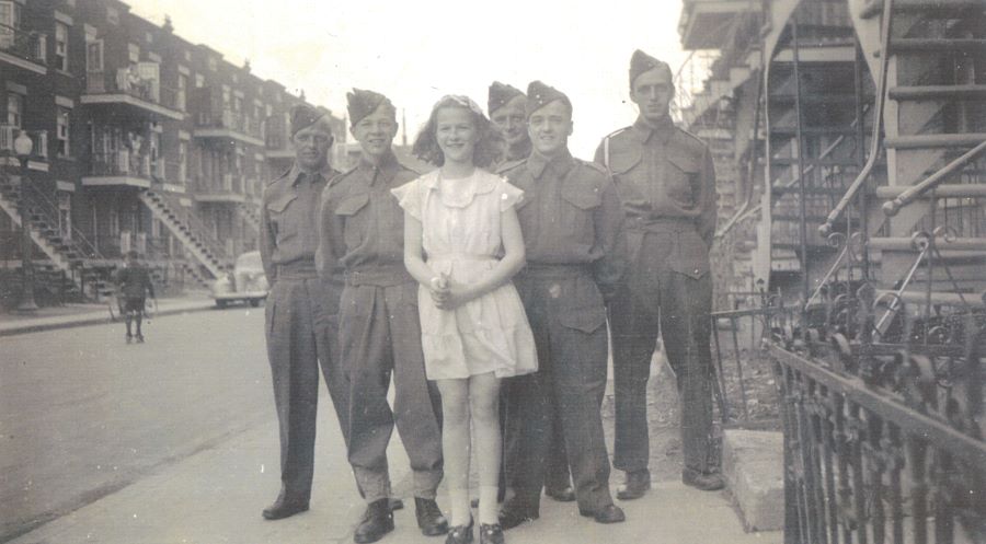Photographie en noir et blanc de 6 personnes debout sur un trottoir, une jeune fille et cinq militaires en uniforme. En arrière-plan, des logements de trois étages avec escaliers et balcons.
