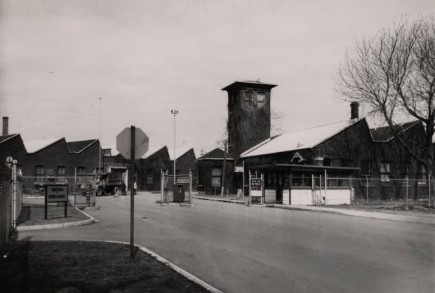 Photographie en noir et blanc de l’entrée routière qui mène aux installations de l’ancienne usine de munitions. L’entrée est munie d’un poste de contrôle et les installations sont clôturées.