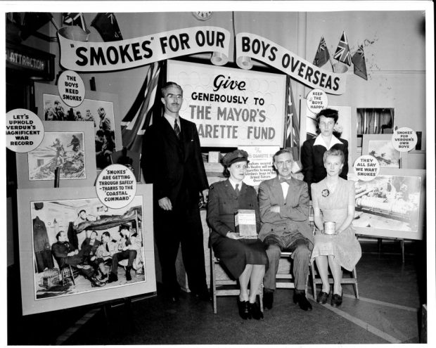 Photographie en noir et blanc montrant deux hommes et trois femmes devant un kiosque promotionnel comportant divers slogans et plusieurs photographies de soldats dans leur quotidien.