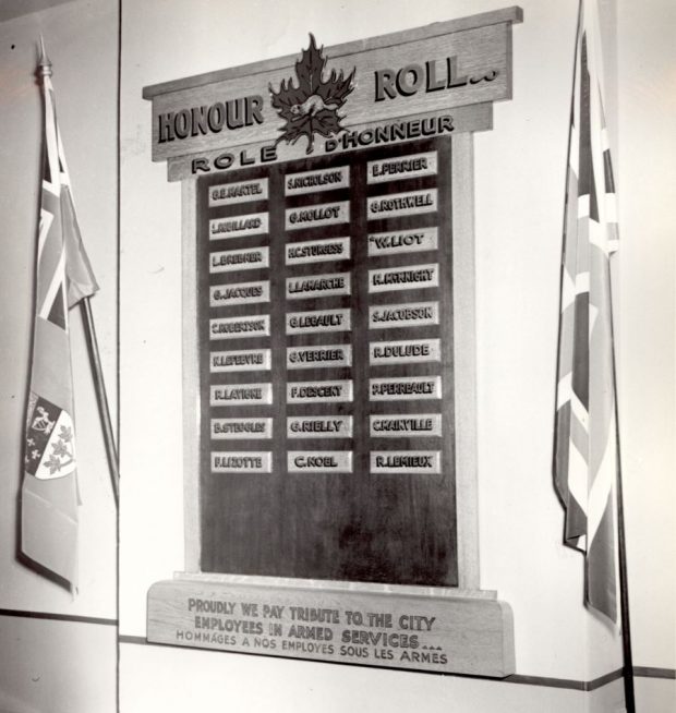 Photographie noir et blanc d’un tableau d’honneur accroché à un mur entre deux drapeaux, un canadien, l’autre britannique. Sur le tableau, 27 petites plaques rectangulaires avec les noms d’employés en service actif. 