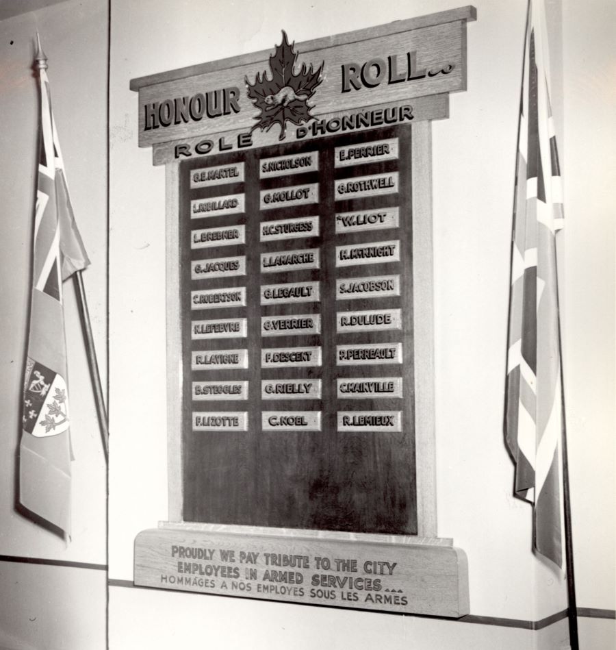 Photographie noir et blanc d’un tableau d’honneur accroché à un mur entre deux drapeaux, un canadien, l’autre britannique. Sur le tableau, 27 petites plaques rectangulaires avec les noms d’employés en service actif. 