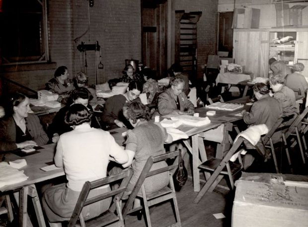 Photographie en noir et blanc de 15 femmes assises à des tables (deux longues rangées) dans un local et coupant des tissus. 