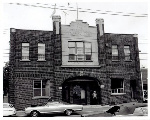  Photographie noir et blanc d’un bâtiment dont la façade indique les inscriptions « The Great War Memorial Hall » et « Lest We Forget ». Un homme se tient devant la porte principale et des voitures sont stationnées près du bâtiment.