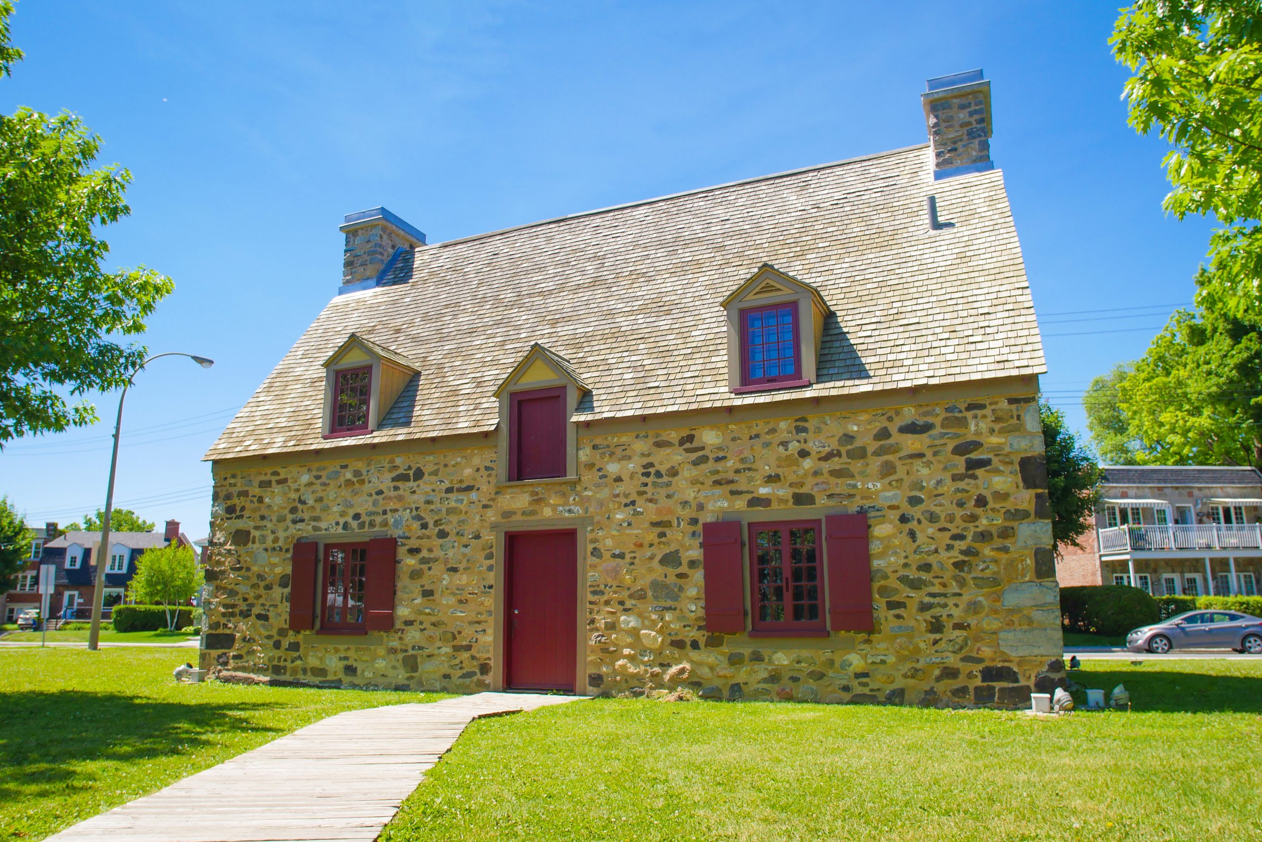 Photographie couleur d'une maison en pierre comportant une porte centrale en bois peinte rouge, deux fenêtres avec volets rouges, deux lucarnes et deux cheminées. En arrière-plan, une rue et deux bâtiments.