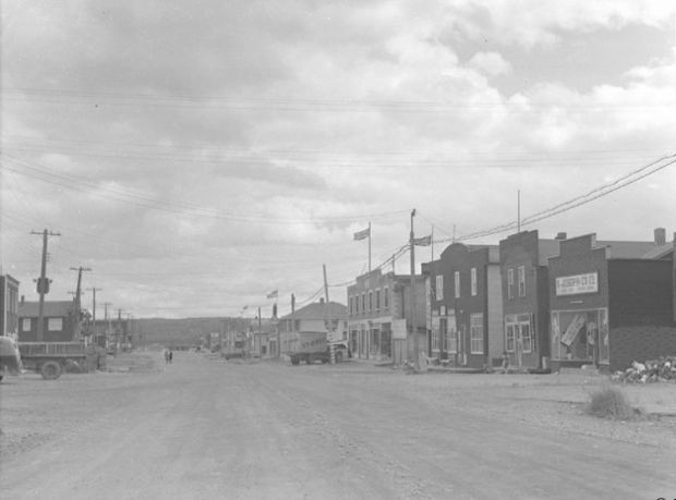 Photographie en noir et blanc d’une route de gravier bordée de bâtiments de construction récente. Plusieurs ont des façades de style Boomtown. Un camion de l’entreprise St-Onge est stationné à droite.