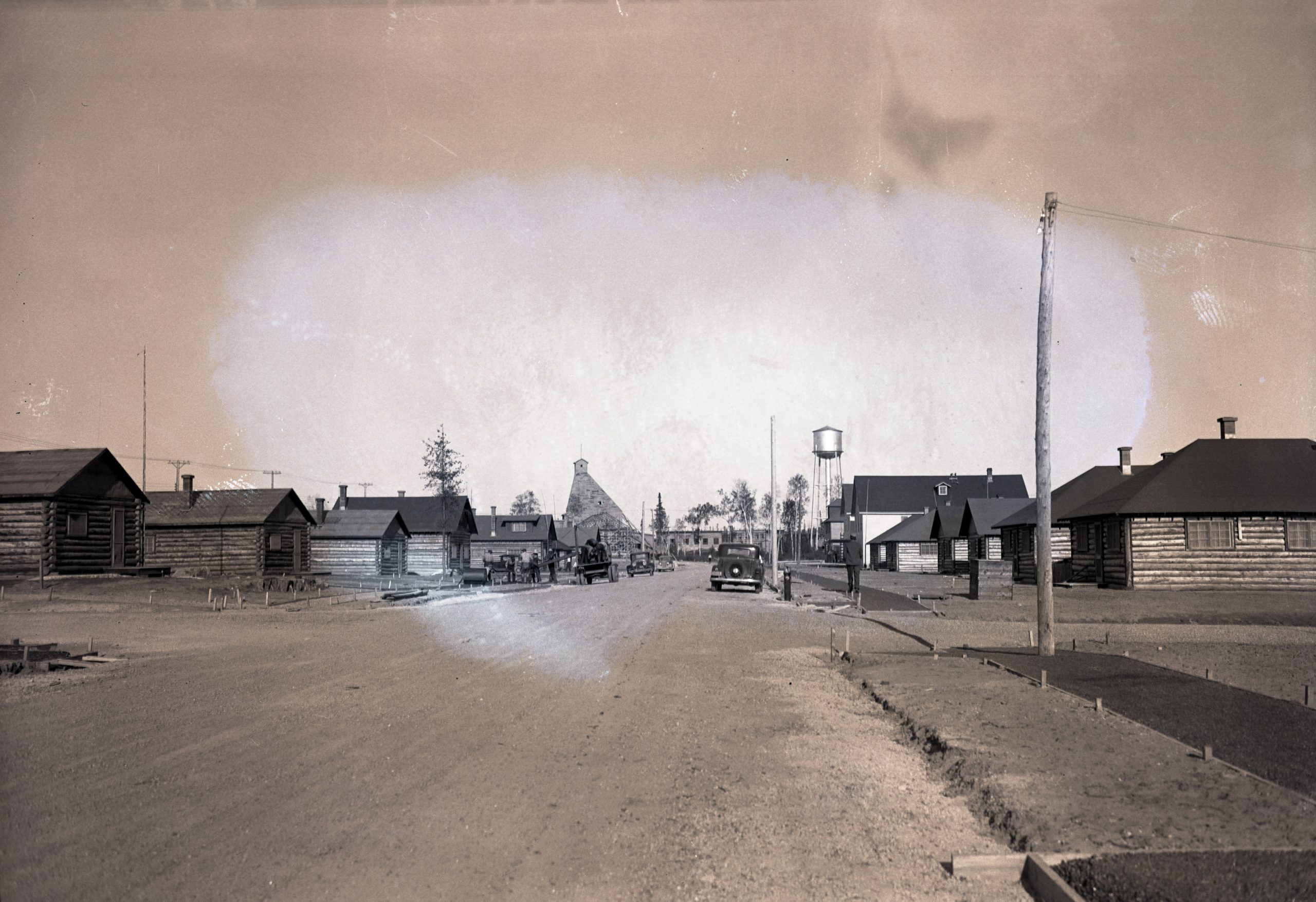 Photographie en noir et blanc d’une rue bordée de cabanes en bois rond. Des hommes travaillent à la construction des trottoirs. En arrière-plan, un château d’eau et un chevalement minier.