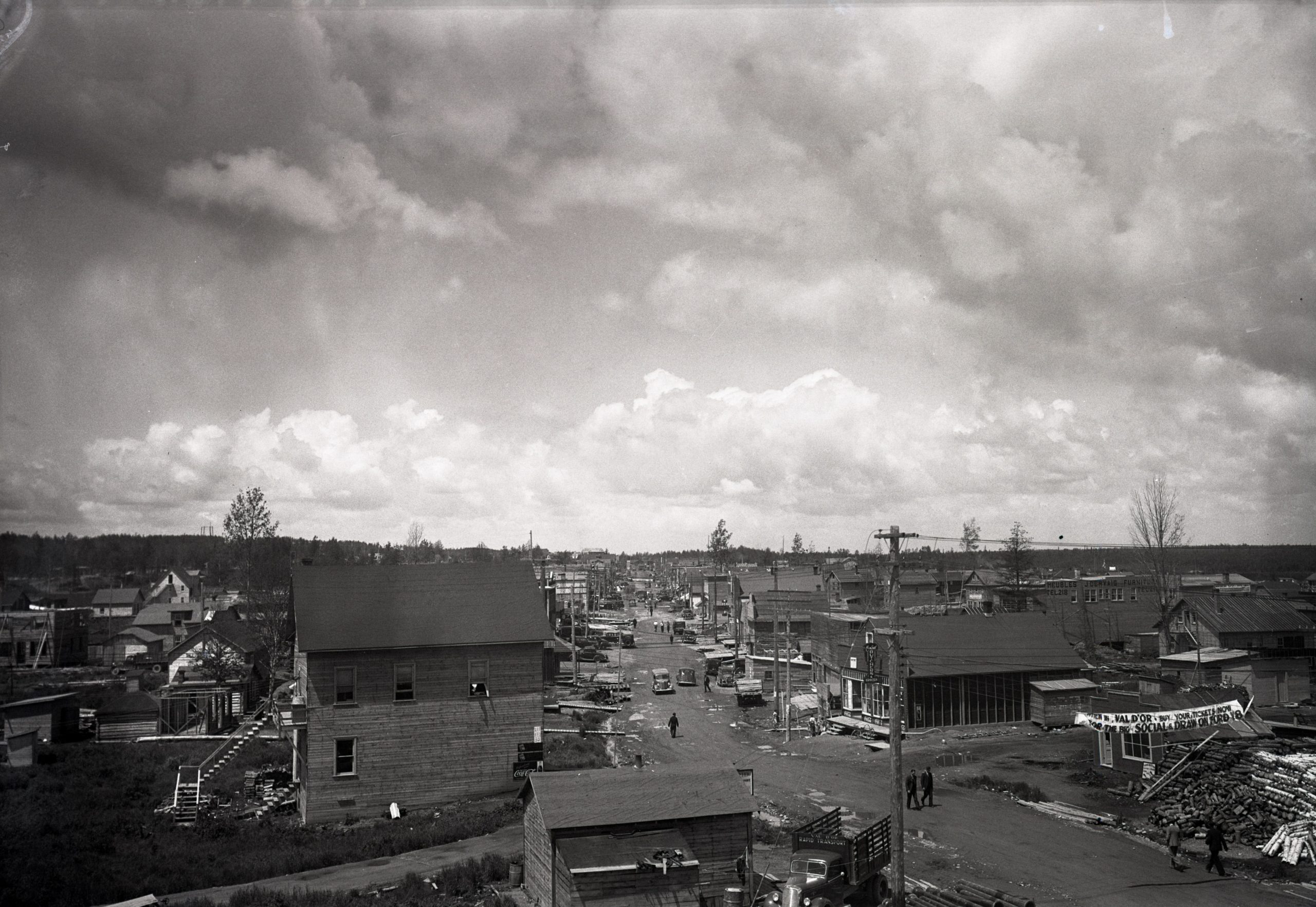 Photographie en noir et blanc d’une rue bordée des deux côtés de bâtiments rudimentaires. Des voitures et des piétons circulent au centre de la route non pavée qui est parsemée de trous d’eau.