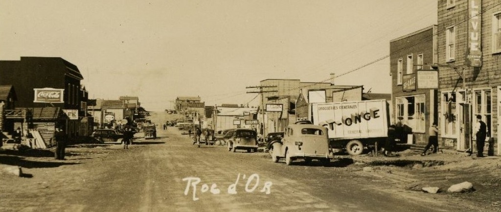 Photographie sépia d’une rue non pavée bordée de bâtiments des deux côtés. Du côté droit, plusieurs voitures et un camion portant l’inscription « St-Onge » sont visibles.