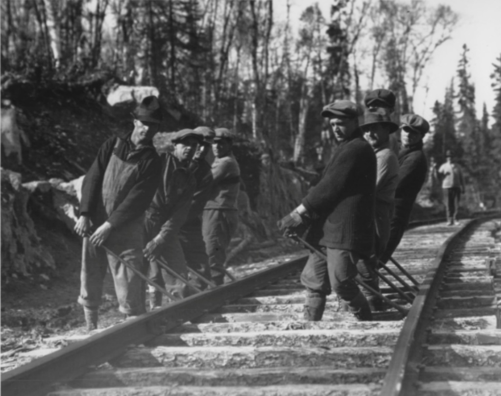 Photographie en noir et blanc d’un groupe de huit hommes qui travaillent sur un chemin de fer avec des barres de fer. À l’arrière-plan, une forêt.