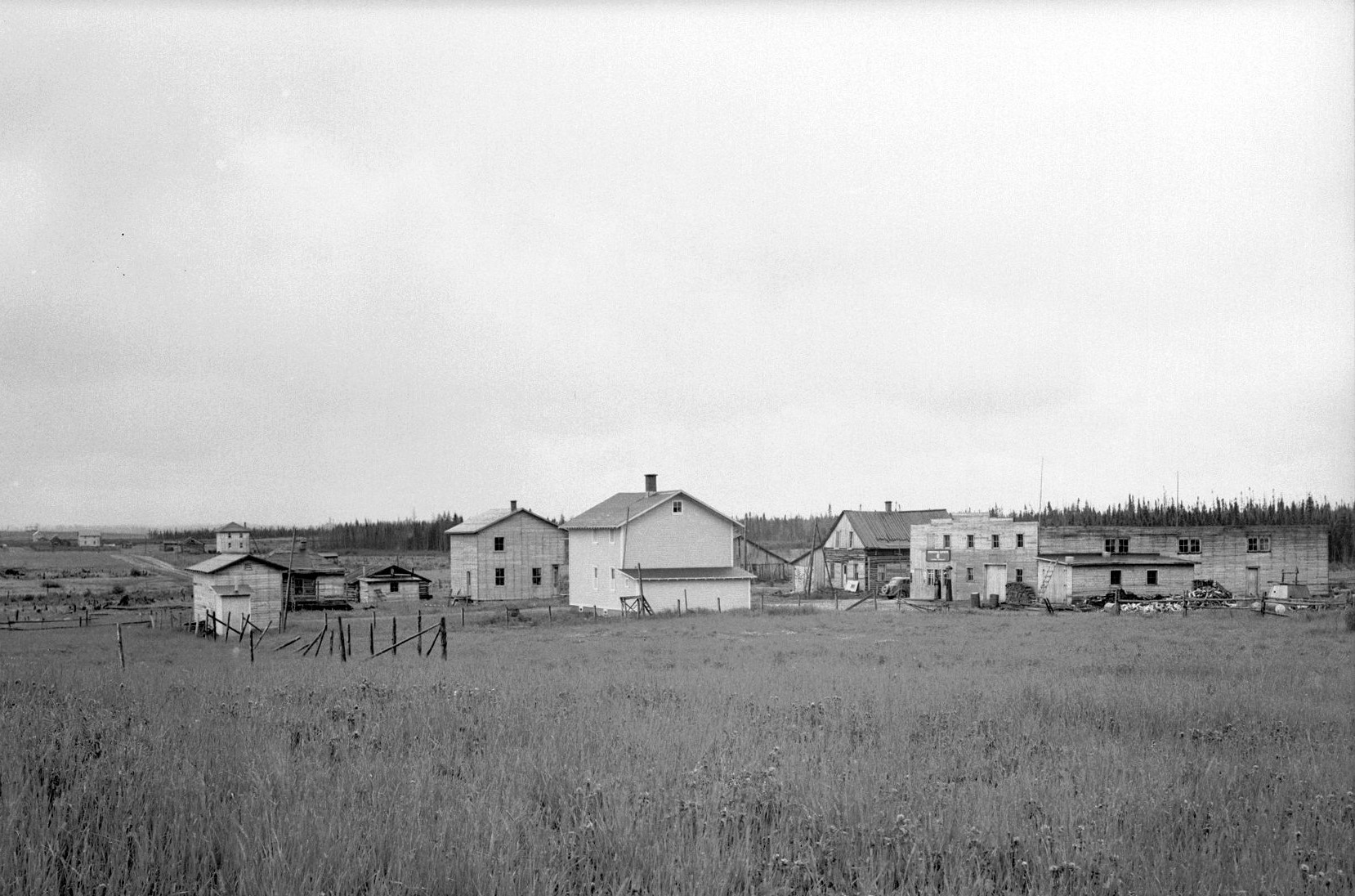 Photographie en noir et blanc d’un hameau d’une dizaine de bâtiments, en bois rond et de planches, entourés d’un champ. À l’arrière-plan, une forêt. 