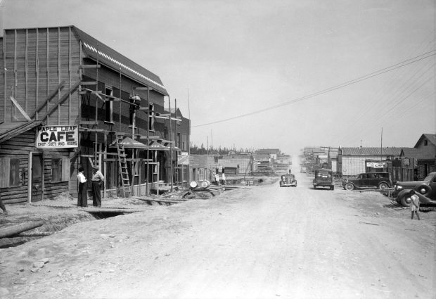 Photographie en noir et blanc d’une route de gravier bordée de bâtiments de planches ou en bois rond. À gauche, le « Maple Leaf Cafe Chop Suey and Rooms » et un immeuble en construction. À droite, plusieurs automobiles et un enfant qui marche. 