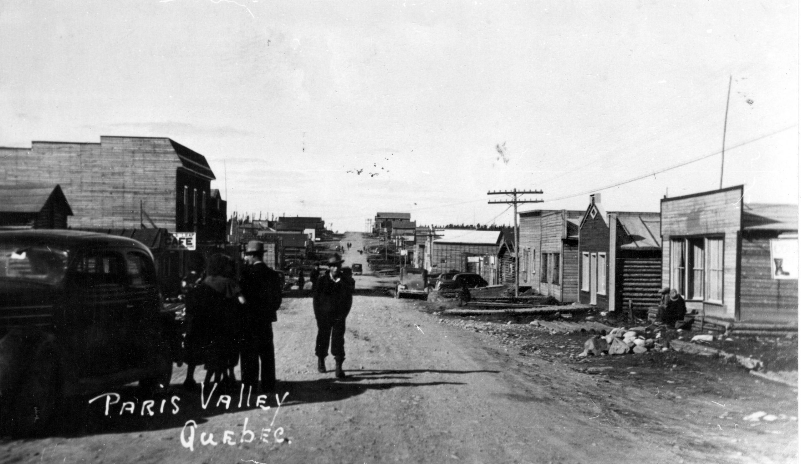 Photographie en noir et blanc d’une route de gravier bordée de bâtiments en bois rond ou de planches. Une automobile et plusieurs personnes sont au milieu de la rue, au centre de la photo. Au bas, l’inscription « Paris Valley Québec. ».